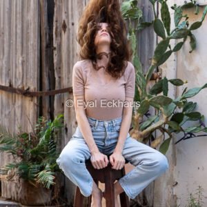 Young woman in jeans sitting on a wooden stool, flipping her long brown hair up against a rustic wooden door and cactus.